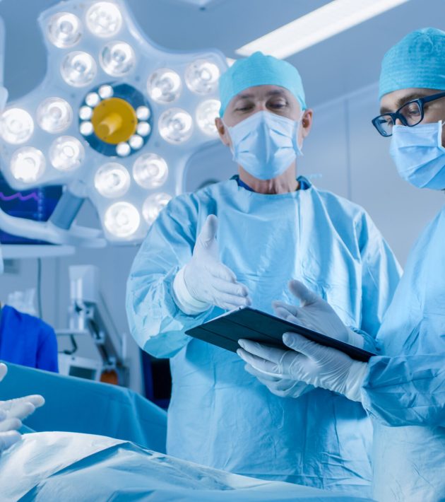 Professional Surgeons and Assistants Talk and Use Digital Tablet Computer During Surgery. They Work in the Modern Hospital Operating Room. Professional Surgeons and Assistants Talk and Use Digital Tablet Computer During Surgery. They Work in the Modern Hospital Operating Room.